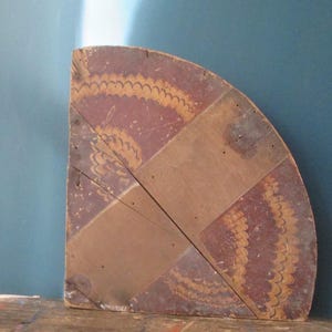 May include: A quarter-circle wooden decorative piece with a geometric design. The piece is primarily brown with accents of red and yellow. The design features a repeating pattern of curved lines, creating a vintage aesthetic.
