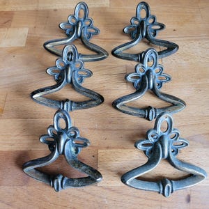 May include: Six antique brass drawer pulls with a decorative floral design. The pulls are shaped like a teardrop with a loop handle.