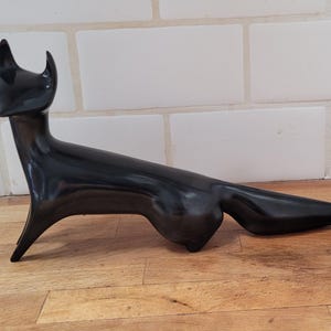 May include: A sleek, black cat sculpture with a long, slender body and pointed ears. The cat is posed in a reclining position, with its tail extended. The sculpture is made of a glossy material and sits on a wooden surface.