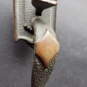 May include: An antique bronze door handle with an ornate, textured design. The handle has a rectangular backplate with a decorative top, a textured grip, and a copper-coloured accent. A vintage hardware piece.