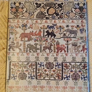 May include: A rectangular decorative item with a detailed, colourful embroidered design. The design features various animals, floral patterns, and geometric shapes in shades of red, green, blue, and brown on a cream background.
