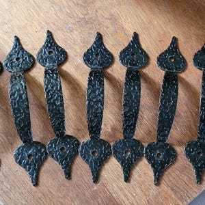 May include: Seven black, textured metal door pulls. Each has a decorative, teardrop-shaped top and bottom, connected by a curved handle. The pulls are arranged on a wooden surface, with a rustic appearance.