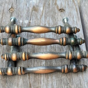 May include: Four metal drawer pulls with a weathered copper and dark bronze finish. Each handle features a central rounded grip, tapered ends, and decorative ribbed detailing near the mounting points, with visible silver-toned screws attached.