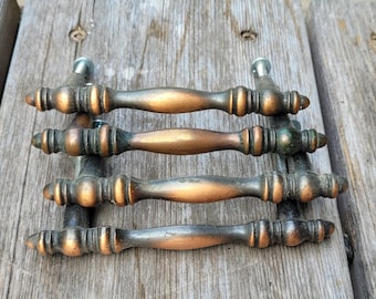 May include: Four metal drawer pulls with a weathered copper and dark bronze finish. Each handle features a central rounded grip, tapered ends, and decorative ribbed detailing near the mounting points, with visible silver-toned screws attached.