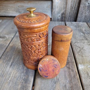 May include: Two wooden containers with intricate carvings. The larger container has a brass knob on the lid. The smaller container is plain. Both containers are sitting on a wooden surface.