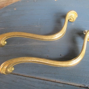 May include: Two gold-toned metal cabinet pulls with a decorative, curved design. The pulls have a smooth, polished finish.
