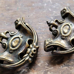 May include: Two antique bronze-colored decorative corner pieces. Each piece has an ornate design with floral and oval accents. The pieces are made of metal and are likely for furniture or architectural use.