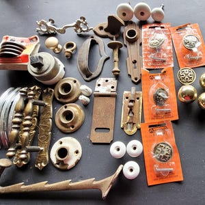 May include: A collection of vintage hardware components, including door knobs, handles, and latches. The assortment features various styles, colors, and materials, such as brass, ceramic, and metal. Some items are new in packaging, while others show signs of age.