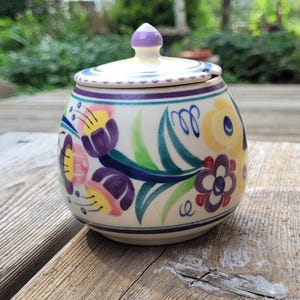 May include: A round ceramic sugar bowl with a matching lid featuring a hand-painted floral pattern in shades of purple, yellow, green, and blue on a cream-colored background. The lid has a small purple knob and a notch for a spoon.
