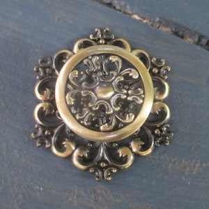May include: A gold-toned metal decorative knob with a floral design. The knob has a raised centre with a circular pattern and is surrounded by a decorative border.