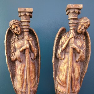 May include: Two gold-painted angel figurines holding columns. Each angel has wings and is holding a column with a decorative top.