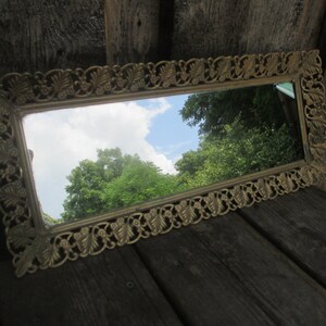 May include: A gold-coloured metal tray with a decorative, ornate design. The tray has a mirror surface that reflects a view of trees and a cloudy sky.