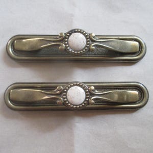 May include: Two antique-style cabinet door pulls with a brass finish and white ceramic knobs. Each knob has a decorative beaded border.