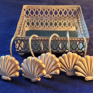 May include: A decorative bathroom set featuring a silver metal tray with a mirrored base and six seashell-shaped hooks. The hooks and tray have a gold and silver finish, set against a dark blue background.
