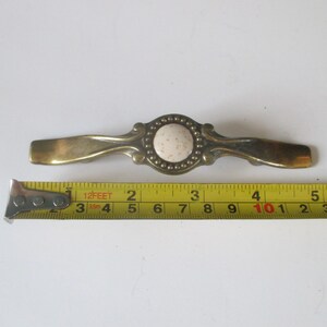May include: A vintage brass drawer pull with a central circular design featuring a speckled, off-white insert surrounded by small, raised dots. The pull measures approximately 5 inches in length, as indicated by the measuring tape.