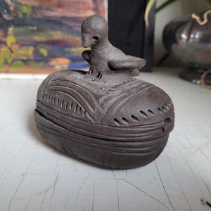 May include: A dark brown ceramic trinket box with a bird-shaped lid. The box has intricate carvings and a smooth, matte finish.