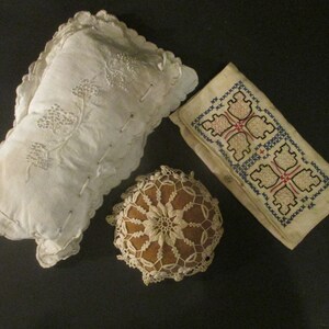May include: Three vintage white fabric items: a pillow with embroidered floral designs, a small crocheted doily, and a rectangular piece with embroidered geometric patterns in blue, red, and white.