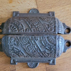 May include: Two antique, cast iron drawer pulls with ornate floral and geometric patterns. The pulls are a weathered grey colour and have mounting holes on each end. The hardware is likely for a cabinet or chest.