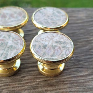 Peut inclure: Quatre boutons de meuble dorés avec des dessus ronds et à motifs. Les dessus présentent un mélange de couleurs vertes, roses et blanches. Les boutons sont disposés en carré, mettant en valeur leur design décoratif.