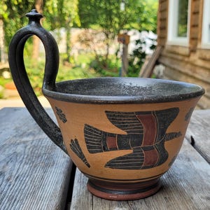 May include: A terracotta-colored ceramic cup with a tall, arched black handle. The exterior features a hand-painted black and dark red bird motif with linear details. The interior rim is black, and the base is a dark, rounded pedestal design.