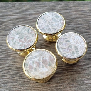 May include: Four cabinet knobs with a gold finish and a circular, patterned top. The tops feature a design with shades of green, pink, and cream. The knobs are arranged on a wooden surface.