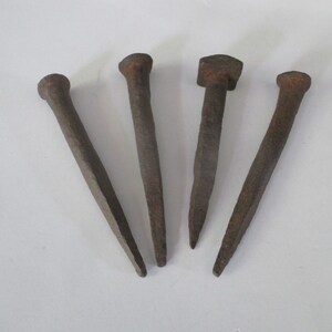 May include: Four antique, rusty metal nails with varying head shapes. The nails are a dark brown color and appear to be hand-forged. The heads include a rounded, a square, and a flat top.
