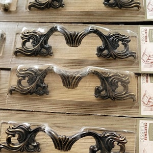 Peut inclure: Tirants de tiroir ornés de couleur bronze dans un emballage transparent. Les poignées ont un design incurvé avec des volutes décoratives et un motif floral central. L'emballage est sur une surface en bois.