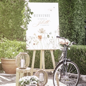 May include: A white sign with the text "Bienvenue au bapt&ecirc;me de Juliette 07 septembre 2023" in French, with a floral design featuring orange, pink, and green flowers. The sign is on an easel, and there is a bicycle and lanterns in the background.