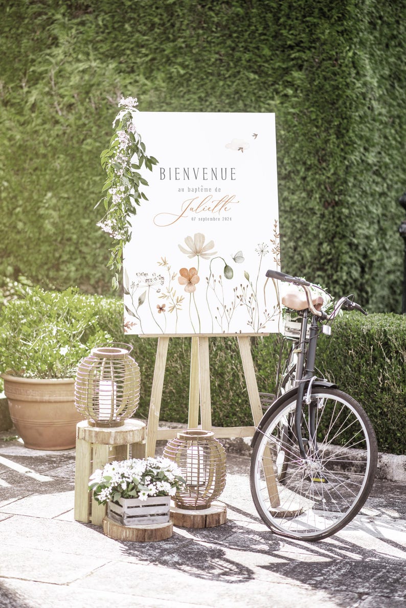 May include: A white sign with the text "Bienvenue au bapt&ecirc;me de Juliette 07 septembre 2023" in French, with a floral design featuring orange, pink, and green flowers. The sign is on an easel, and there is a bicycle and lanterns in the background.