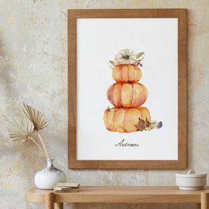 May include: A watercolor painting of a stack of three pumpkins with a white flower on top. The pumpkins are orange and the flower is white with a brown center. The painting is framed in a wooden frame and the word "Automne" is written below the pumpkins.
