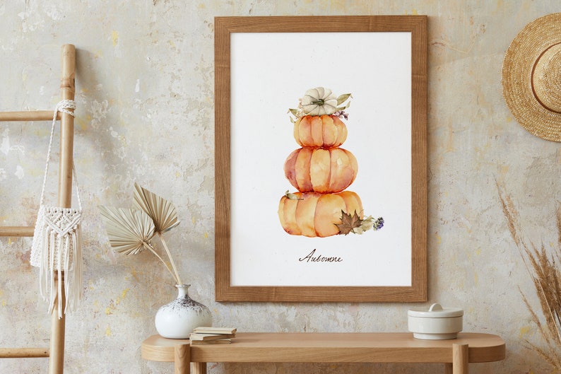 May include: A watercolor painting of a stack of three pumpkins with a white flower on top. The pumpkins are orange and the flower is white with a brown center. The painting is framed in a wooden frame and the word "Automne" is written below the pumpkins.