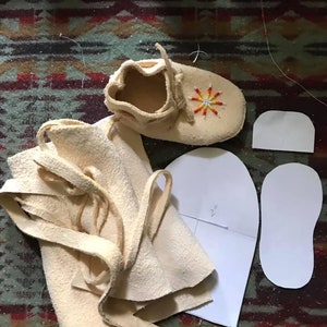 May include: A close-up of a pair of unfinished leather moccasins with a beaded sun design. The moccasins are laid out on a patterned rug with a piece of paper with a pattern for the moccasins. The paper has the text "2" on it.