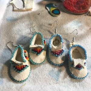 May include: Four pairs of handmade white moccasin earrings with blue and colorful bead accents. The earrings have a small, shoe-like design with beaded details around the edges and on the front. The background is a white surface with craft supplies.
