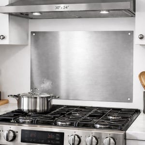 May include: A stainless steel range hood with two lights is mounted above a gas range. A stainless steel backsplash is mounted on the wall. A stainless steel pot with a lid is on the stove. Kitchen utensils and bottles are on the countertop.