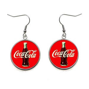 May include: A pair of silver earrings with red round pendants featuring a Coca-Cola bottle and logo.