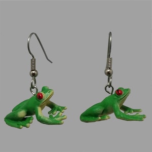 May include: A pair of green frog earrings with red eyes. The frog charms are detailed with visible legs and arms, and are suspended from silver-colored fish hook ear wires. The earrings are set against a neutral gray background.