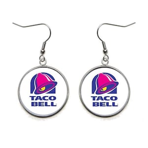 May include: A pair of silver earrings with a white background and the Taco Bell logo, featuring a pink bell with a blue outline and the text "Taco Bell" in blue.