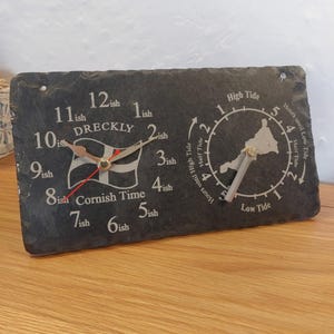 May include: A slate clock with two clock faces. One shows the time with the words "Cornish Time" and "Dreckly". The other shows the tide times with a map of Cornwall. The clock has silver and red hands.