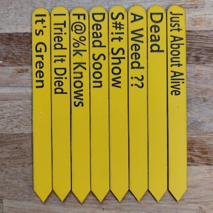 May include: Eight yellow wooden plant markers with black text. The text on the markers reads: "Just About Alive", "Dead", "A Weed??", "S#!t Show", "Dead Soon", "F@%k Knows", "I Tried It Died", and "It's Green".