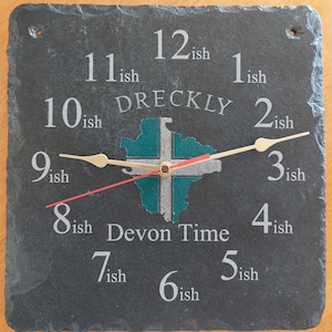 May include: A black slate clock with gold hands and a green and white map of Devon, England. The clock face reads "12ish, 11ish, 10ish, 9ish, 8ish, 7ish, 6ish, 5ish, 4ish, 3ish, 2ish, 1ish" and "DRECKLY" and "Devon Time".