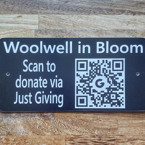 May include: A black rectangular sign with white text. It reads "Woolwell in Bloom" at the top, with "Scan to donate via Just Giving" below. A QR code is on the right. The sign has two holes for mounting.