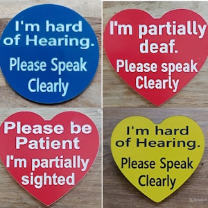 May include: Four heart-shaped and round stickers with different messages about hearing and sight impairments. The stickers are red, blue, yellow, and white. The text on the stickers reads: "I'm hard of hearing. Please speak clearly", "I'm partially deaf. Please speak clearly", "Please be patient. I'm partially sighted", and "I'm hard of hearing. Please speak clearly".