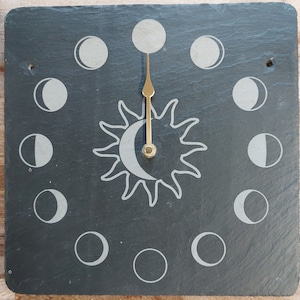 May include: A square, dark grey slate clock with a sun and crescent moon design at the centre. The clock face displays the phases of the moon around the edge. The clock hands are gold-coloured.