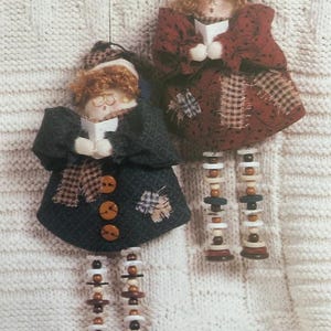 May include: Two hanging fabric dolls with curly ginger hair, one wearing a dark charcoal coat and the other a deep red coat. Both dolls hold white books and feature long, dangling legs constructed from stacked brown, white, and blue buttons.