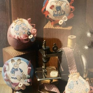 May include: Three round fabric pincushions featuring embroidered rabbit figures, a small teardrop-shaped fabric ornament, a miniature black metal sewing machine, a spool of thread, various loose buttons, and metal scissors on a wooden surface.