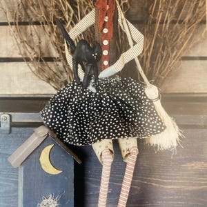 May include: A fabric doll with orange hair wearing a black hat, a brown vest with white buttons, and a black skirt with white stars. The doll holds a black cat and a broom, and wears striped leggings with black boots. A blue wooden house with a yellow moon sits nearby.