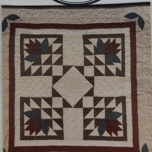 May include: A square wall hanging quilt featuring a Bear's Paw Baskets #117 pattern in shades of tan, brown, and dark blue. The design includes a central diamond, four corner blocks with floral motifs, and text reading DeCuyper Trading Co. 27" Wallhanging.