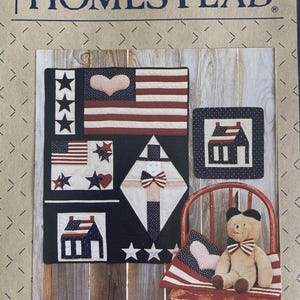 May include: A Liberty Homestead craft pattern featuring a quilt design with American flag motifs, stars, hearts, and house shapes. A tan teddy bear with a striped bow tie sits on a red wooden chair next to a small square quilt with a house graphic.