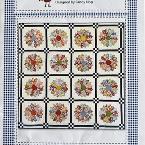 May include: A sewing pattern titled American Jane Patterns Miss Muffet's Tuffet designed by Sandy Klop. The cover features a quilt design with sixteen floral star-shaped blocks arranged in a grid, separated by dark blue and white checkered sashing.
