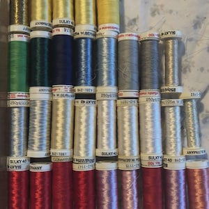 May include: A collection of twenty-eight assorted spools of sewing thread arranged in four rows. The spools feature various colors including gold, green, blue, yellow, silver, grey, white, red, and pink, with labels reading Sulky and Gütermann.